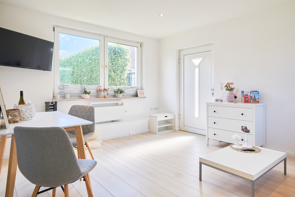 A well-lit living space features a large window allowing natural light to fill the room. A simple white table is paired with two grey chairs. A white sideboard and a small shelf provide storage, while a wall-mounted TV adds entertainment options.