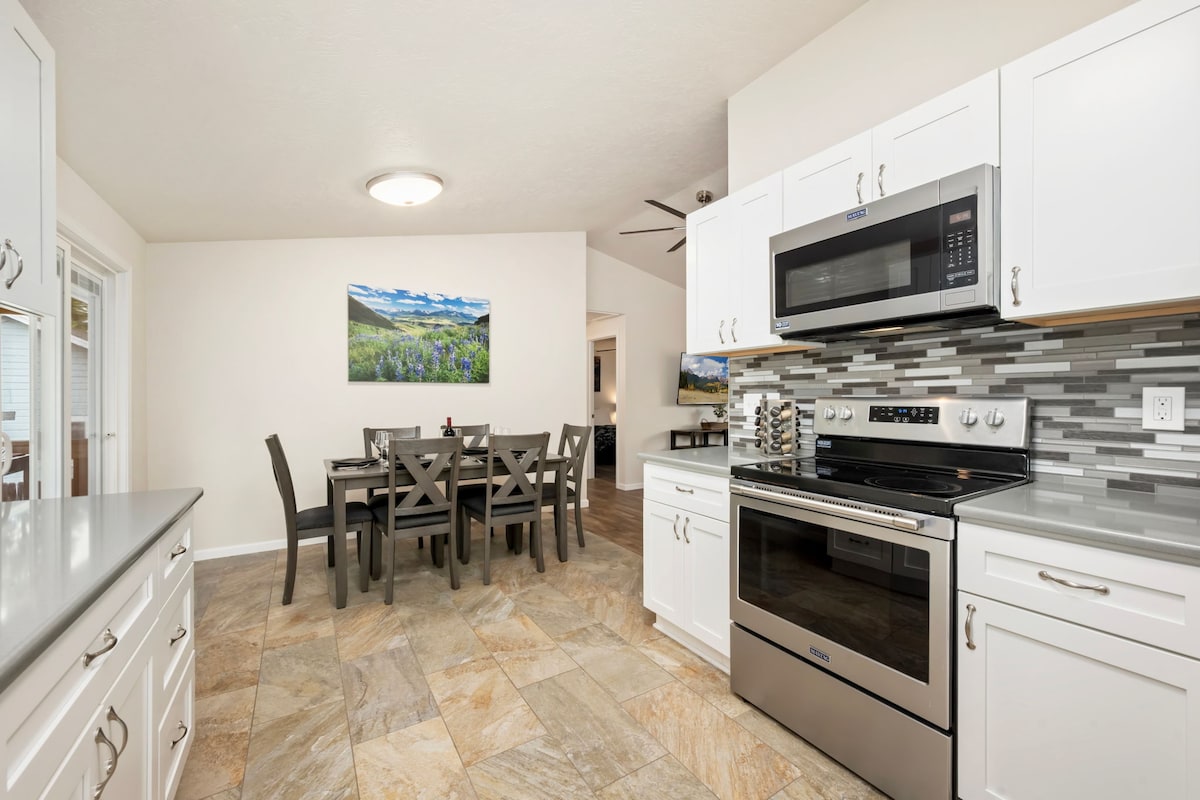 The kitchen features modern appliances, including a stainless steel microwave and oven. Light-colored cabinets complement the neutral tile flooring. A dining area with a large table and chairs is visible in the background, alongside a vibrant mountain-themed artwork on the wall.