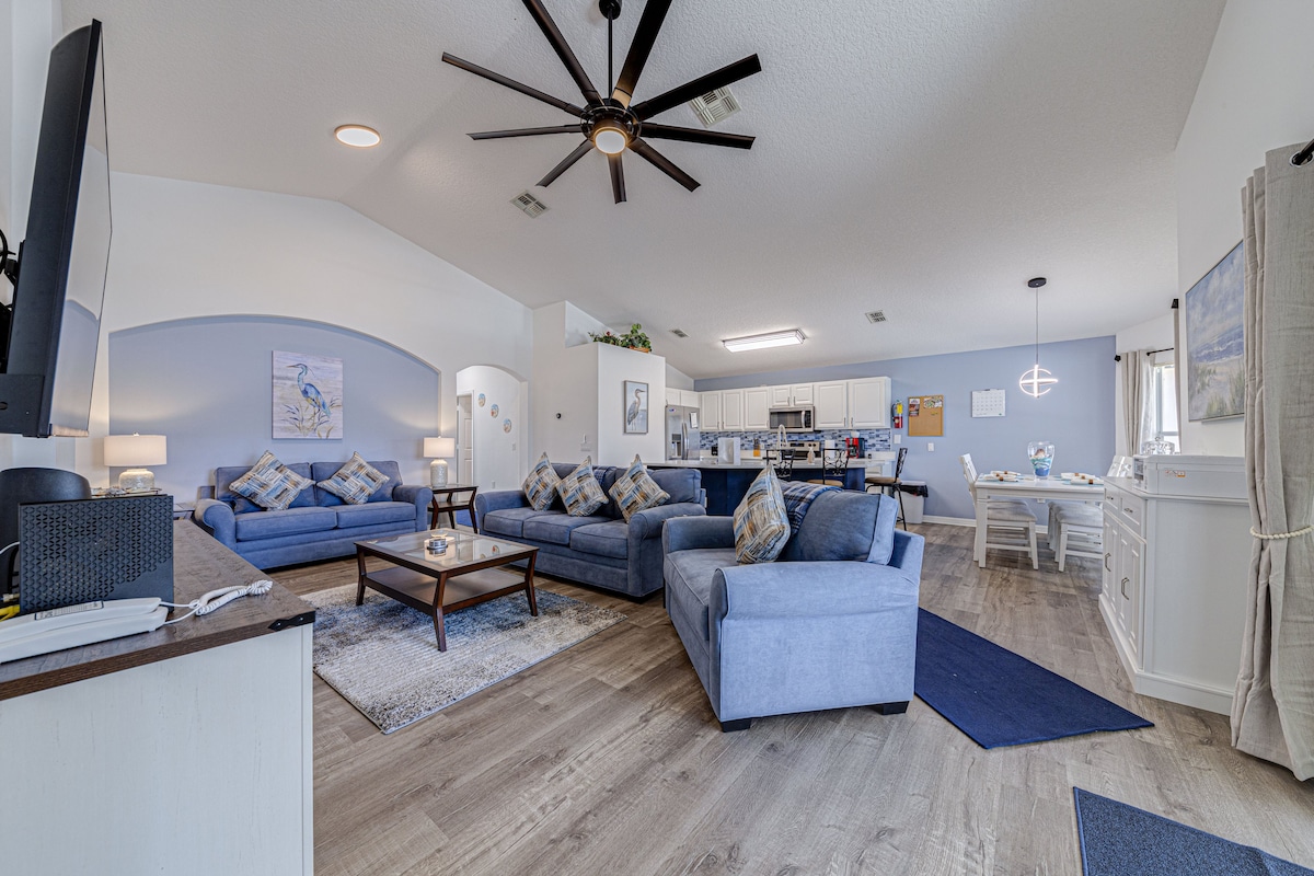 An open-plan living area features comfortable blue seating arranged around a glass coffee table, with decorative cushions enhancing the space. Natural light enters through large windows, illuminating the modern decor. The kitchen is visible in the background, enhancing the welcoming atmosphere.