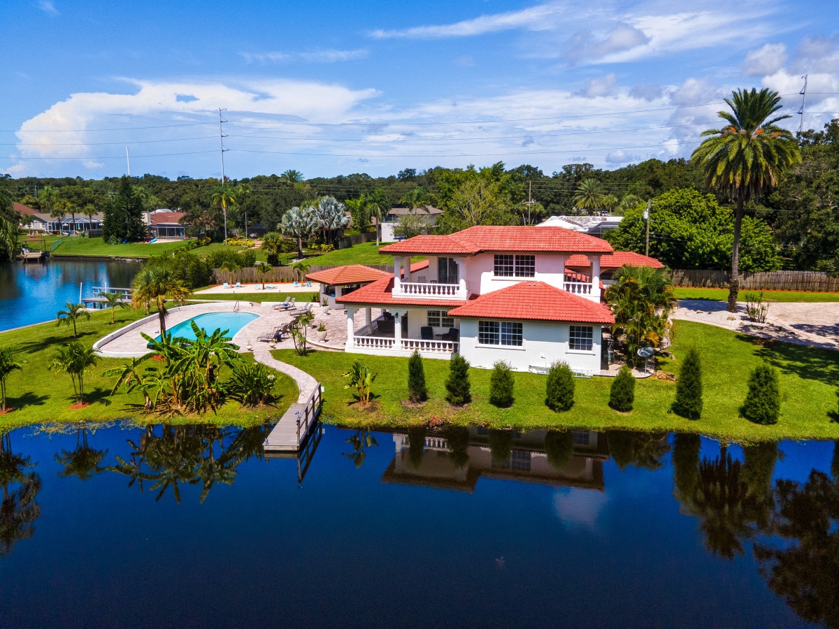 The exterior of The Belleair Estate is showcased from an aerial view, featuring lush landscaping surrounding the home. A private pool glistens alongside the water, with the expansive lawn leading to a private boat dock, creating a serene waterfront setting.