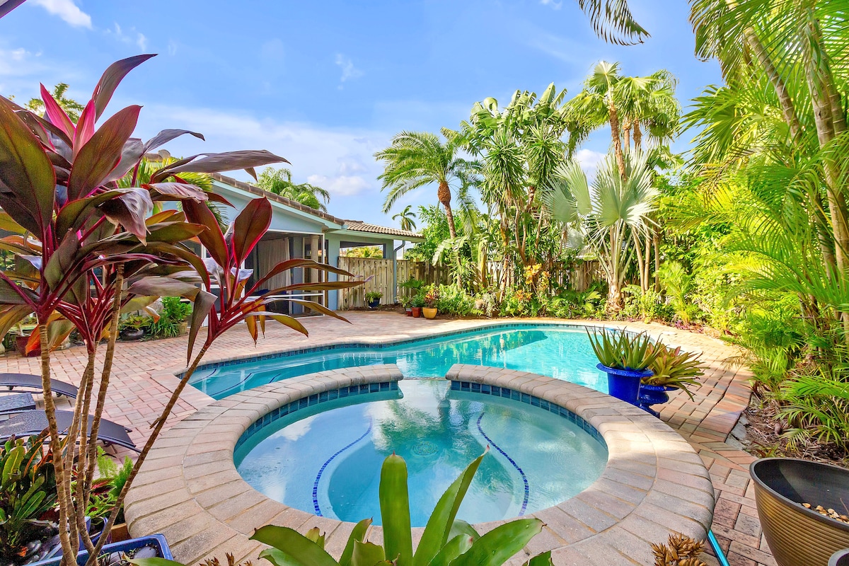 A tropical oasis is showcased, featuring a large pool accompanied by a hot tub, surrounded by lush greenery and vibrant plants. The patio area reflects a welcoming space with additional lounge seating, while a serene atmosphere invites relaxation under the Florida sun.