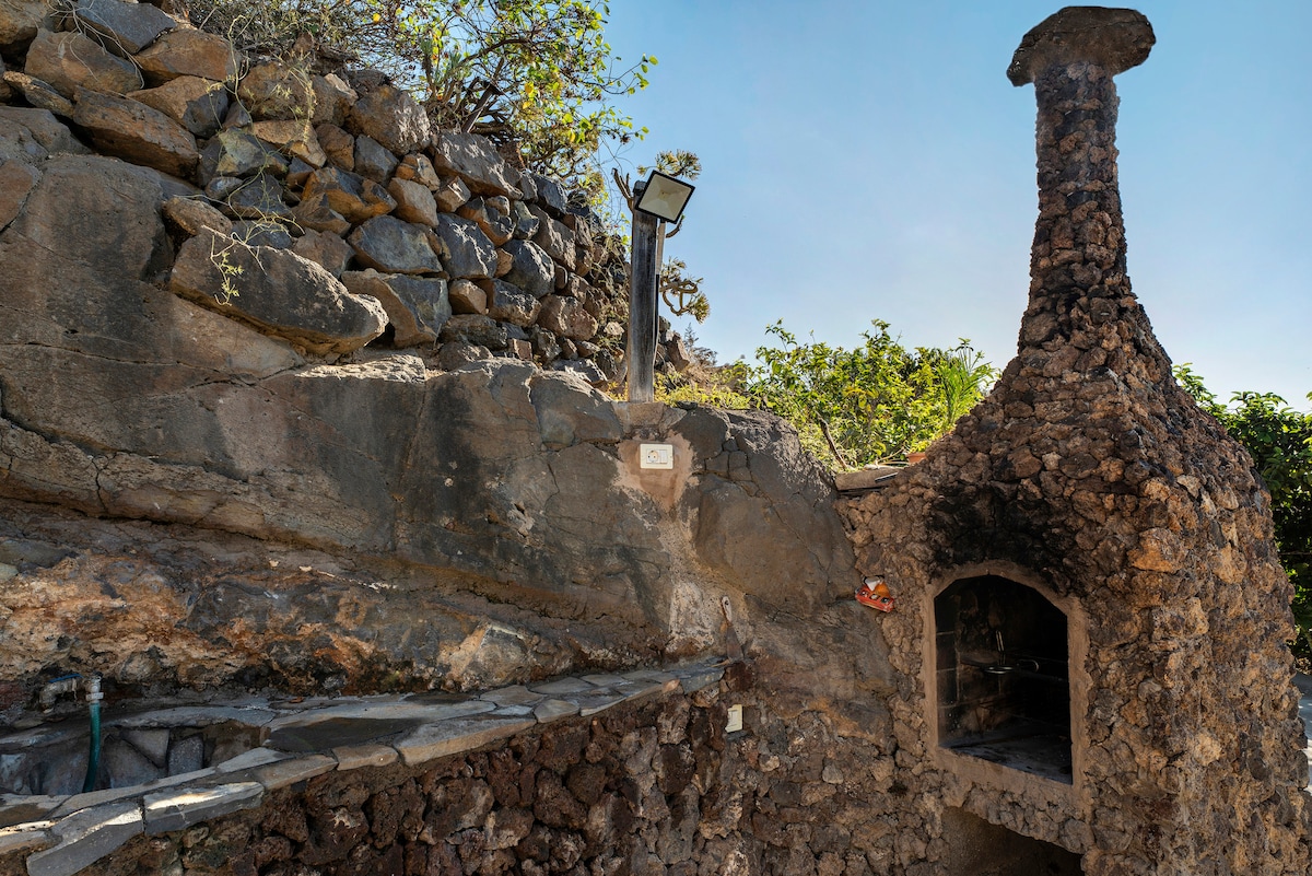 An outdoor barbecue area is nestled against a rocky landscape, featuring a stone-built grill with a tall chimney. Natural greenery frames the space, complemented by a nearby solar-powered light fixture.