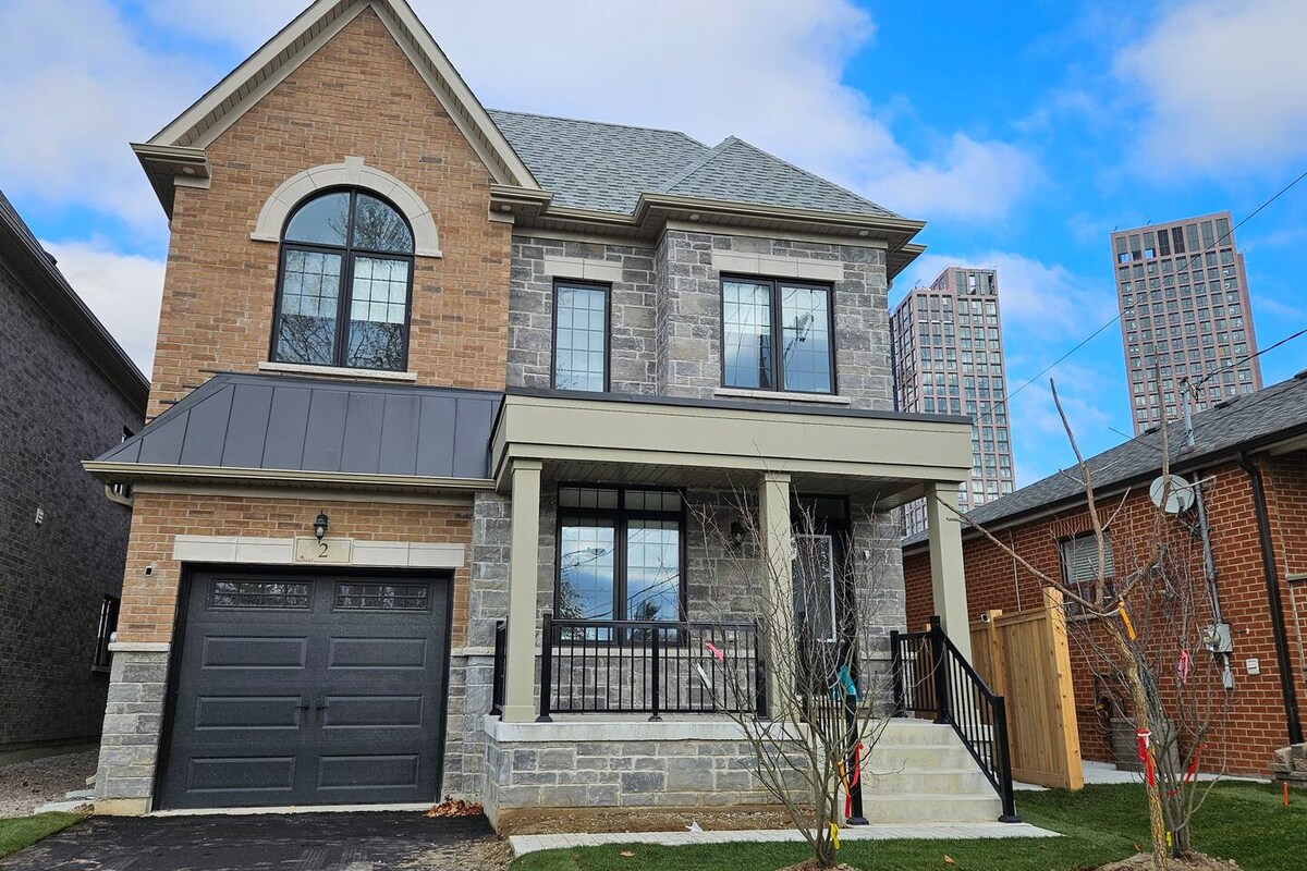 A newly-built detached house features a combination of brick and stone facades. A covered front porch provides access through the central entrance. The driveway accommodates one vehicle, with a modern garage door visible. The backdrop includes surrounding buildings, indicating the central location.