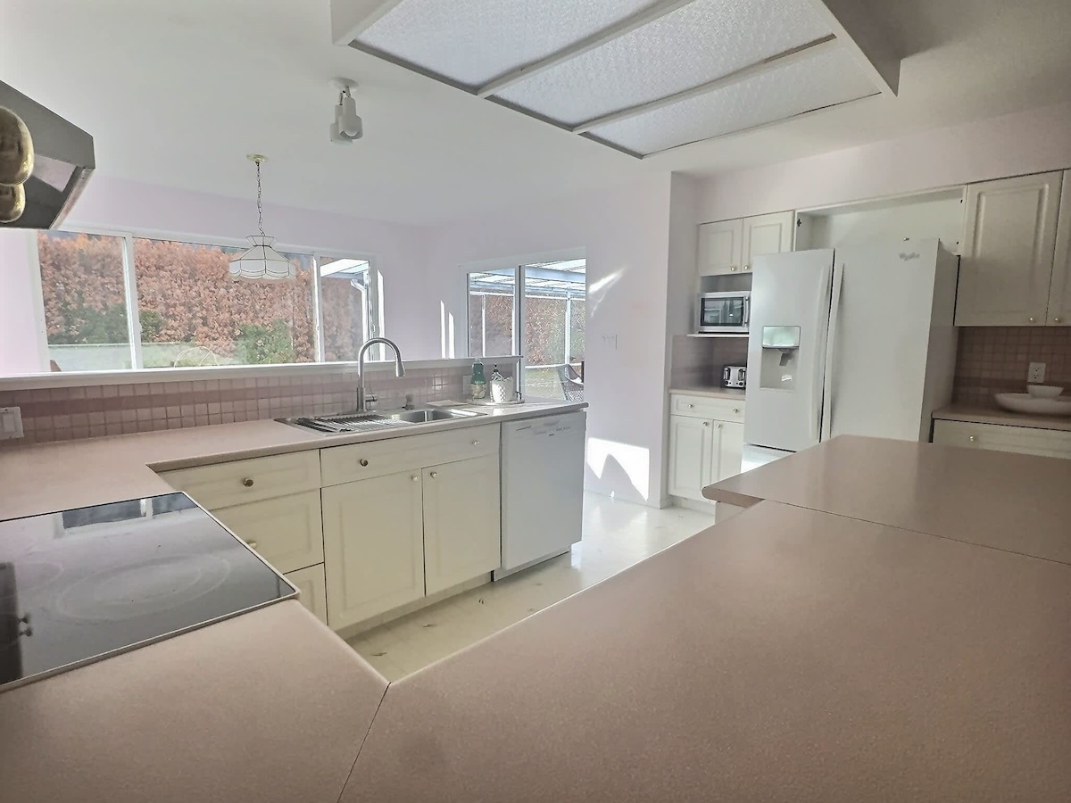 The kitchen features a modern, open layout with white cabinetry and light countertops. Large windows allow ample natural light to illuminate the space, highlighting the sink and cooking area. A refrigerator and microwave are visible, contributing to a functional kitchen setup.