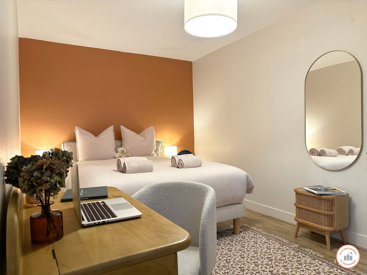 A well-appointed bedroom is visible, featuring a spacious bed with decorative pillows and neatly arranged towels. A mirror reflects soft ambient light. A wooden desk with a laptop sits opposite the bed, complemented by a wicker side table and a decorative rug.