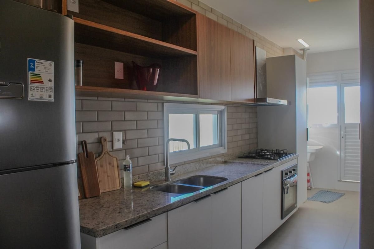 A modern kitchen is equipped with granite countertops and wooden cabinetry. Stainless steel appliances, including a refrigerator and gas stove, are present. Natural light enters through a window, illuminating the space. A cutting board and dish soap are visible on the countertop.