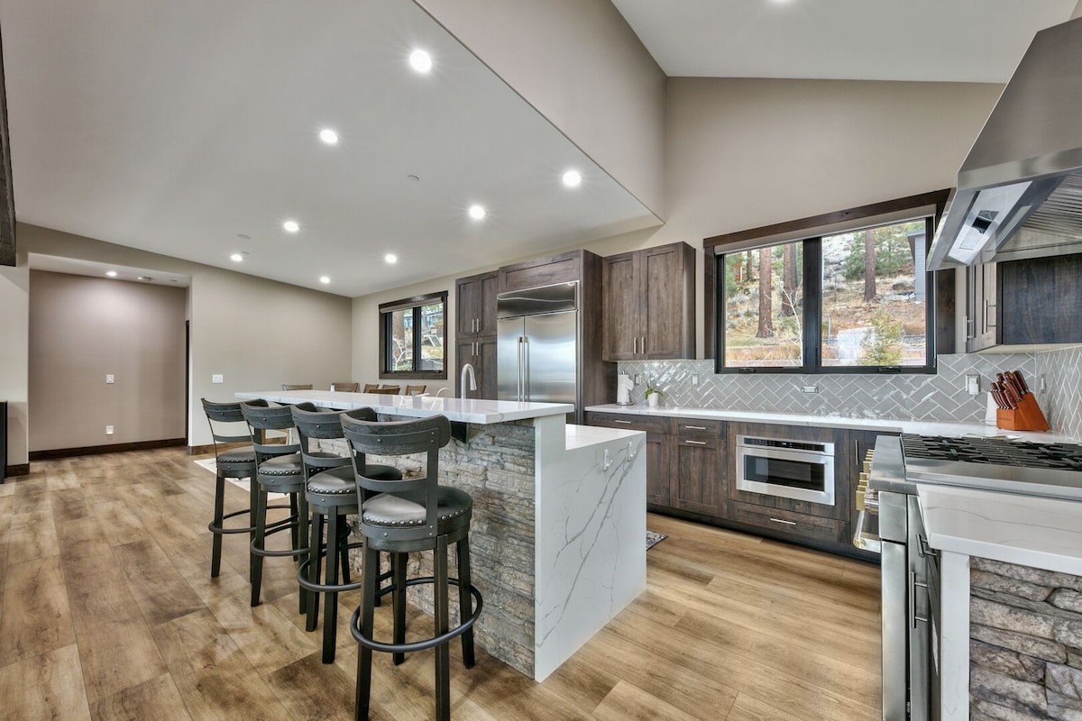 A modern kitchen is displayed, featuring an expansive island with seating for six. Sleek cabinetry and high-end appliances complement the open layout, while large windows offer views of nature. Warm wood flooring enhances the inviting atmosphere throughout the space.
