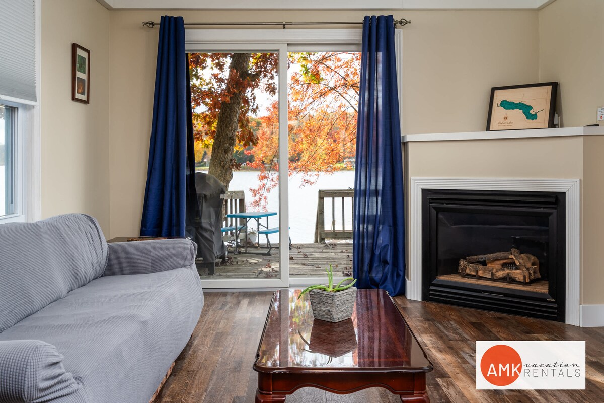 A living area features a gray sofa, a wooden coffee table, and a gas fireplace. Large glass doors provide access to the outdoor deck, framed by vibrant autumn foliage. Dark blue curtains complement the natural light entering the space.