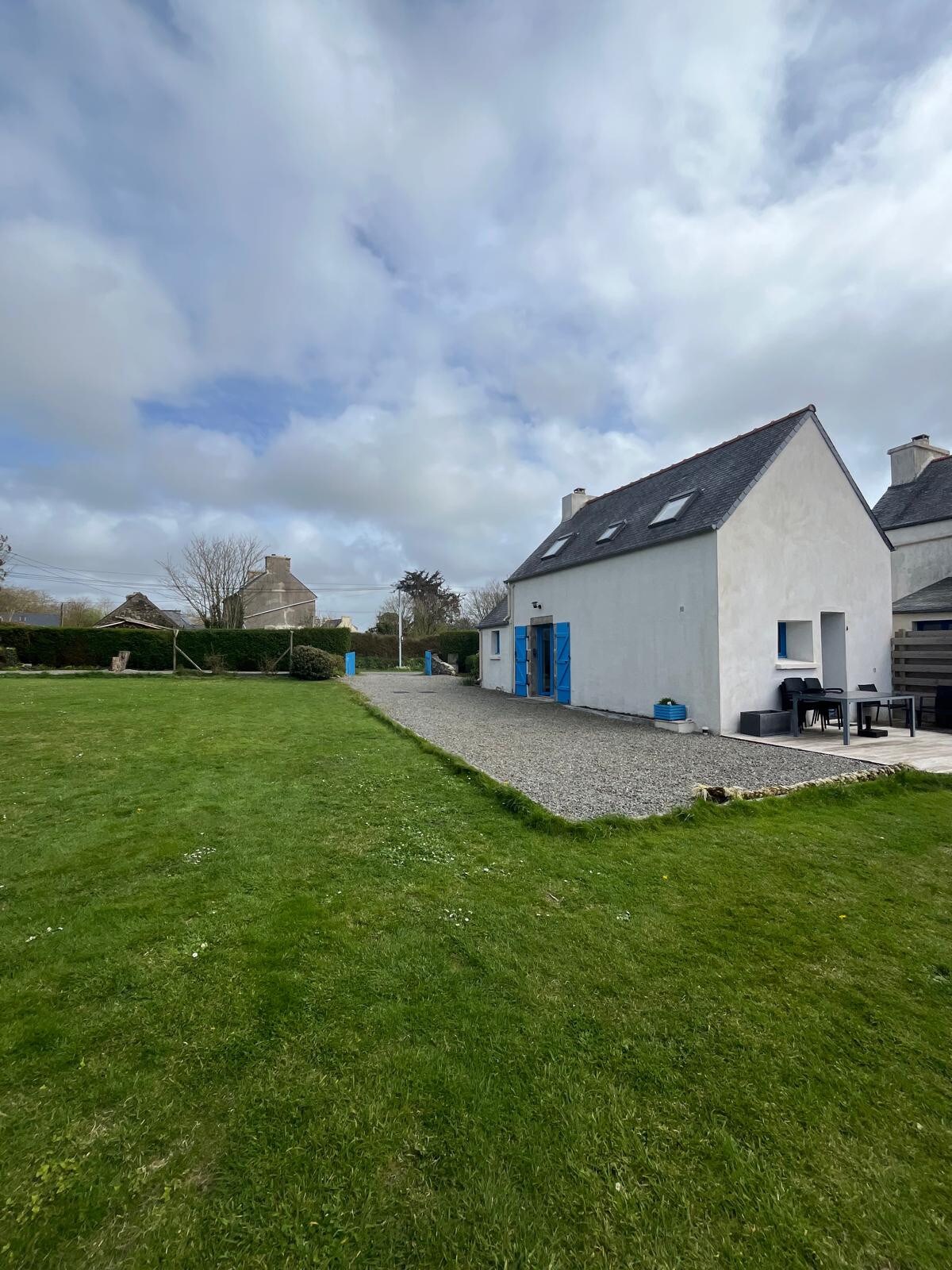 The exterior of a two-story, white house is shown, featuring a sloped roof and blue accents. A spacious grassy area surrounds the home, which has patio space for outdoor seating. The landscape is complemented by scattered clouds in a partially sunny sky.