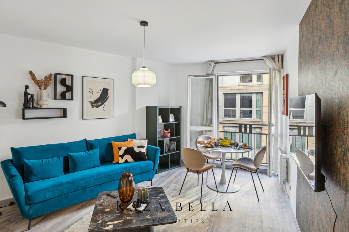 A modern living space features a vibrant blue sofa, accompanied by a wooden coffee table and decorative items. A dining area with a round table and four chairs is positioned near large windows, allowing ample natural light. Artwork adorns the walls, adding character to the room.