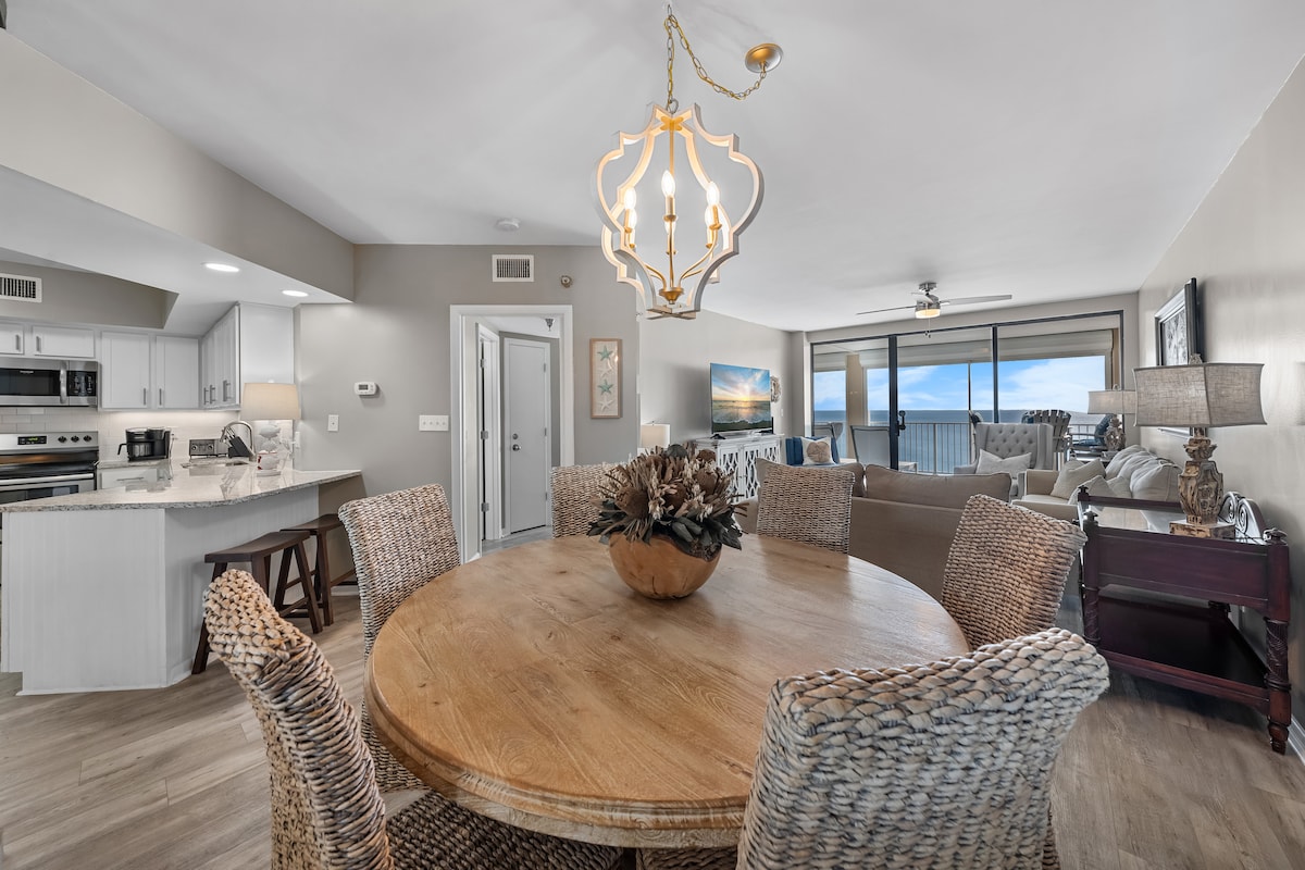 A dining area features a large round wooden table surrounded by six woven chairs. Soft natural light enters through large windows, offering a view of the ocean. In the background, modern furnishings and a chandelier add a touch of elegance to the open layout.