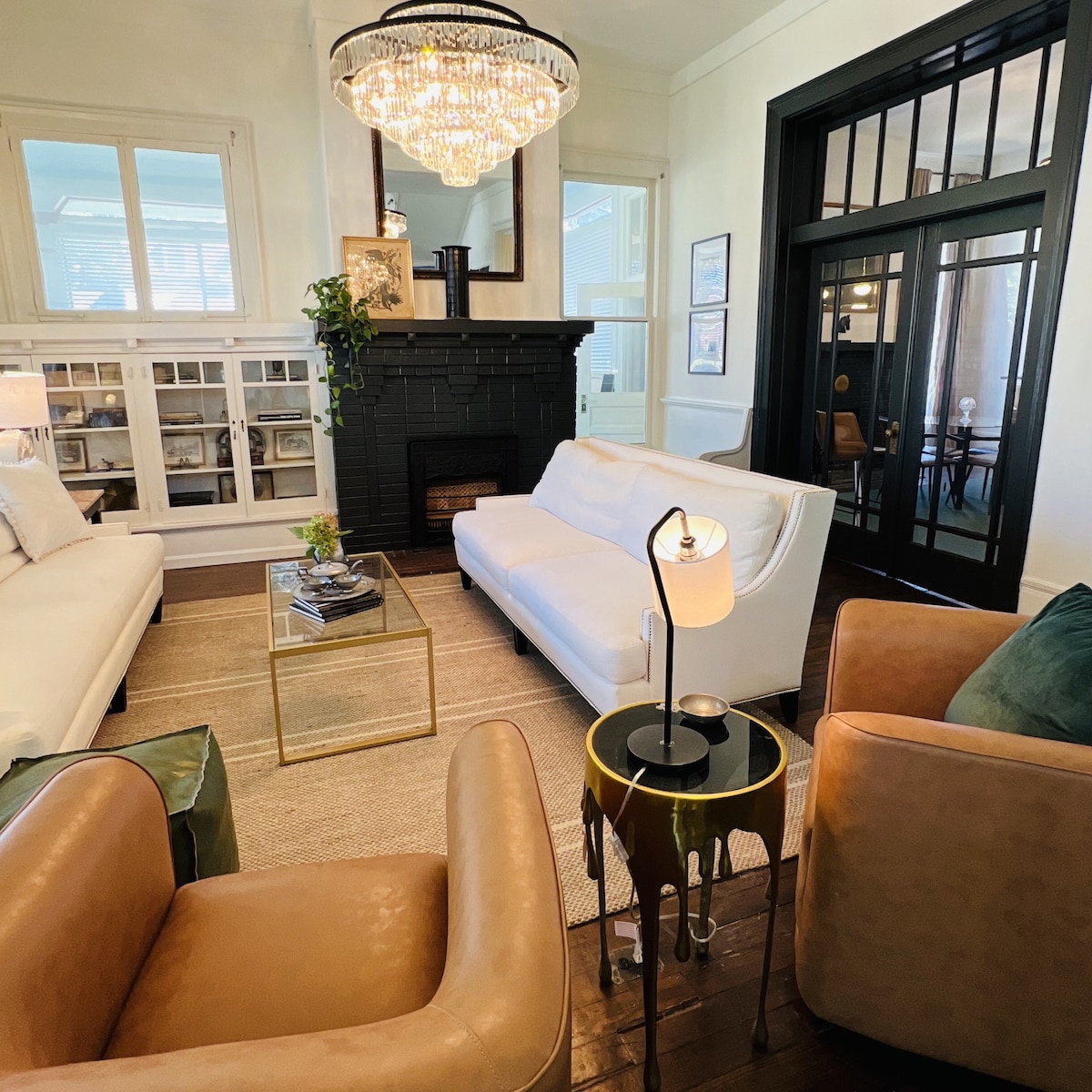The inviting living area features a mix of modern and classic furnishings. A sleek white sofa and two tan armchairs are arranged around a glass coffee table on a natural fiber rug. A chandelier hangs elegantly from the ceiling, and a black fireplace adds a cozy touch.