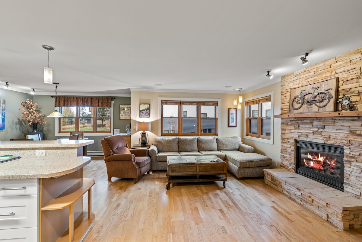 The spacious living area features a large sectional sofa and an armchair, arranged around a coffee table. A stone fireplace adds warmth, while multiple windows enhance natural light. The kitchen area is visible, showcasing a breakfast bar with seating.