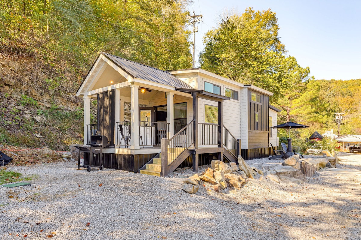 The cabin is situated on a gravel landscape, with a welcoming entrance featuring a deck and steps. A patio invites relaxation, and outdoor furniture can be seen. Surrounding trees provide natural greenery, creating a serene environment.