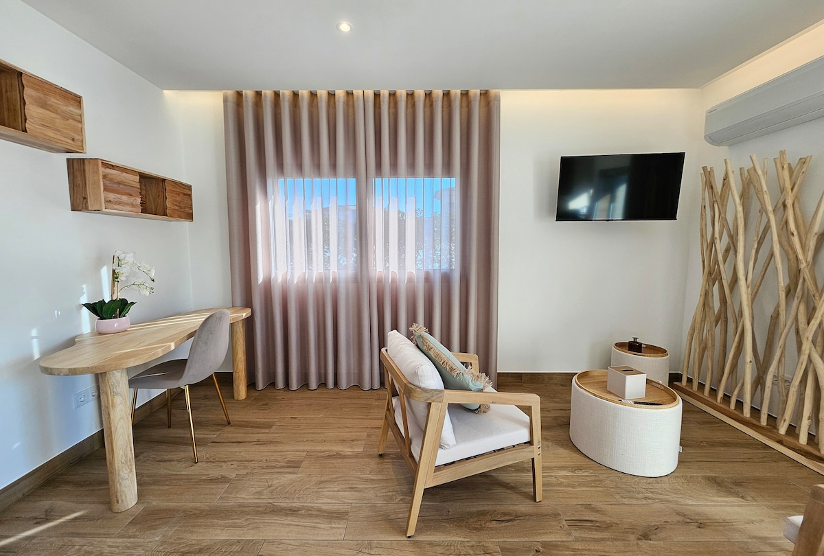 A well-lit living area features large windows dressed with sheer curtains, allowing natural light to filter through. A wooden table and a comfortable chair sit beside a modern coffee table. The decor includes natural materials, creating a calming and elegant space.