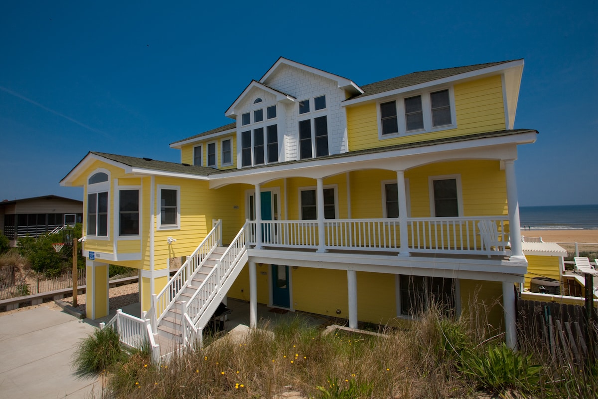 A bright yellow beachfront home features multiple gables and expansive windows. A welcoming front porch with white railings invites entry, while a staircase leads visitors to the upper levels. The sandy beach and sparkling ocean provide a scenic backdrop.
