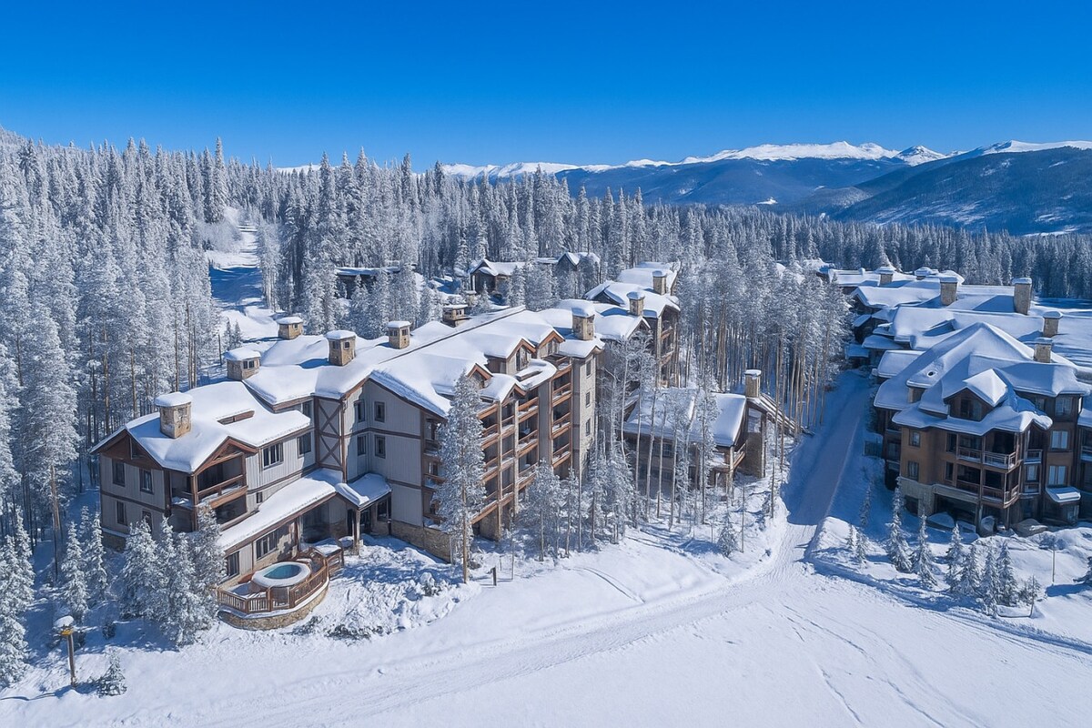 Aerial view of a snow-covered resort, surrounded by a landscape of evergreen trees. The buildings feature sloped roofs blanketed in white snow, with a hot tub visible on a deck. The clear blue sky above complements the wintry scene.