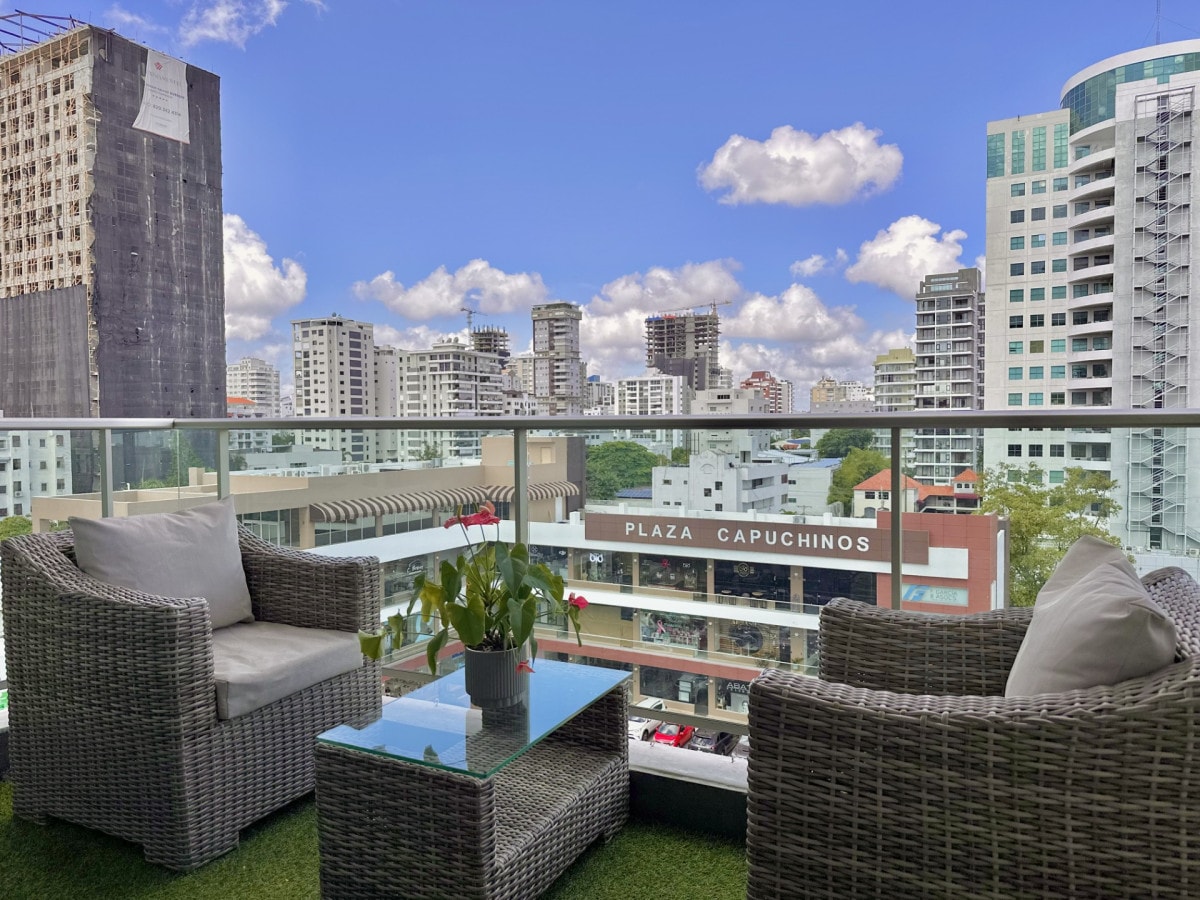 A balcony setup is featured with two woven armchairs and a glass-top table. Potted plants add a touch of greenery. In the background, city buildings rise against a blue sky with fluffy clouds, creating a vibrant urban atmosphere.