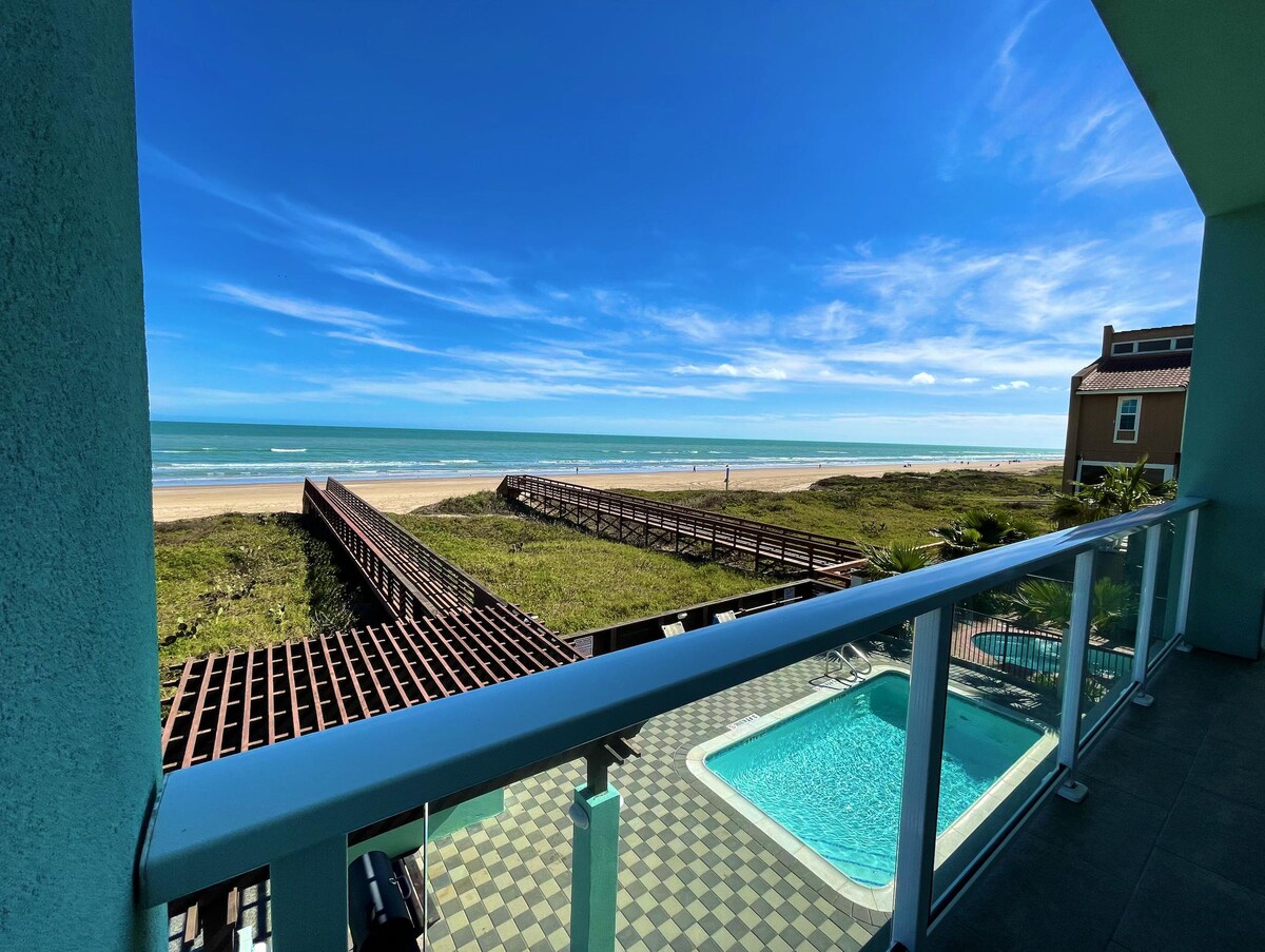 A coastal view is captured from a private balcony, showcasing a sandy beach and the ocean under a clear blue sky. A wooden boardwalk leads to the shore, while a swimming pool can be seen in the foreground, surrounded by landscaped greenery.