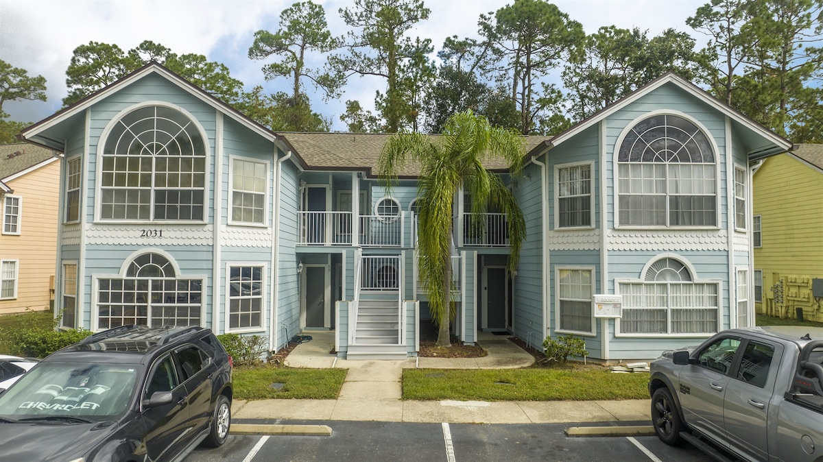 A two-story blue building is presented, featuring large arched windows and white railings along the balconies. Lush green landscaping surrounds the entrance, while parking spaces are visible in front, accommodating multiple vehicles.