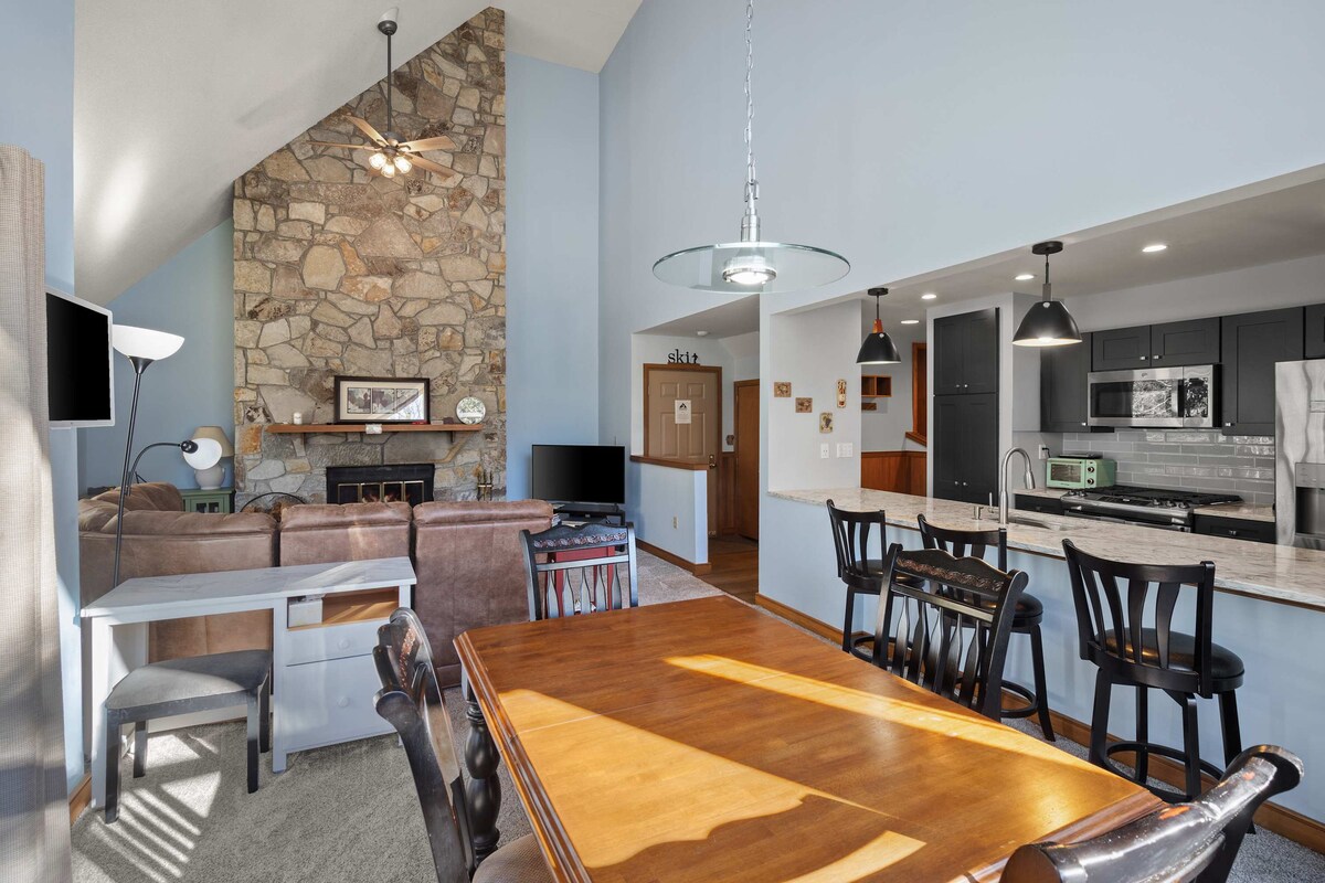 An open dining and living area features a wooden dining table with black chairs and a light source casting shadows on the floor. A stone fireplace serves as the room's focal point, while a cozy couch and a large television are also visible, contributing to a warm setting.