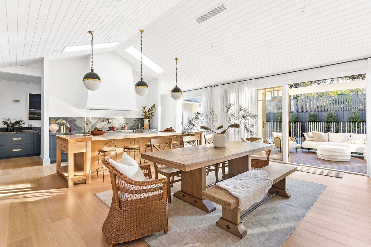 An open-plan living and dining area is showcased, featuring a large wooden dining table surrounded by woven chairs. Natural light filters through sheer curtains, illuminating the space. A spacious kitchen with pendant lights and bar seating is visible in the background, with sliding doors leading to an outdoor terrace.