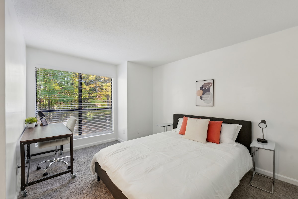 A spacious bedroom features a large bed with a white comforter accented by colorful pillows. A desk with a chair sits nearby, illuminated by natural light streaming through a large window with blinds. A minimalist art piece adorns the wall.