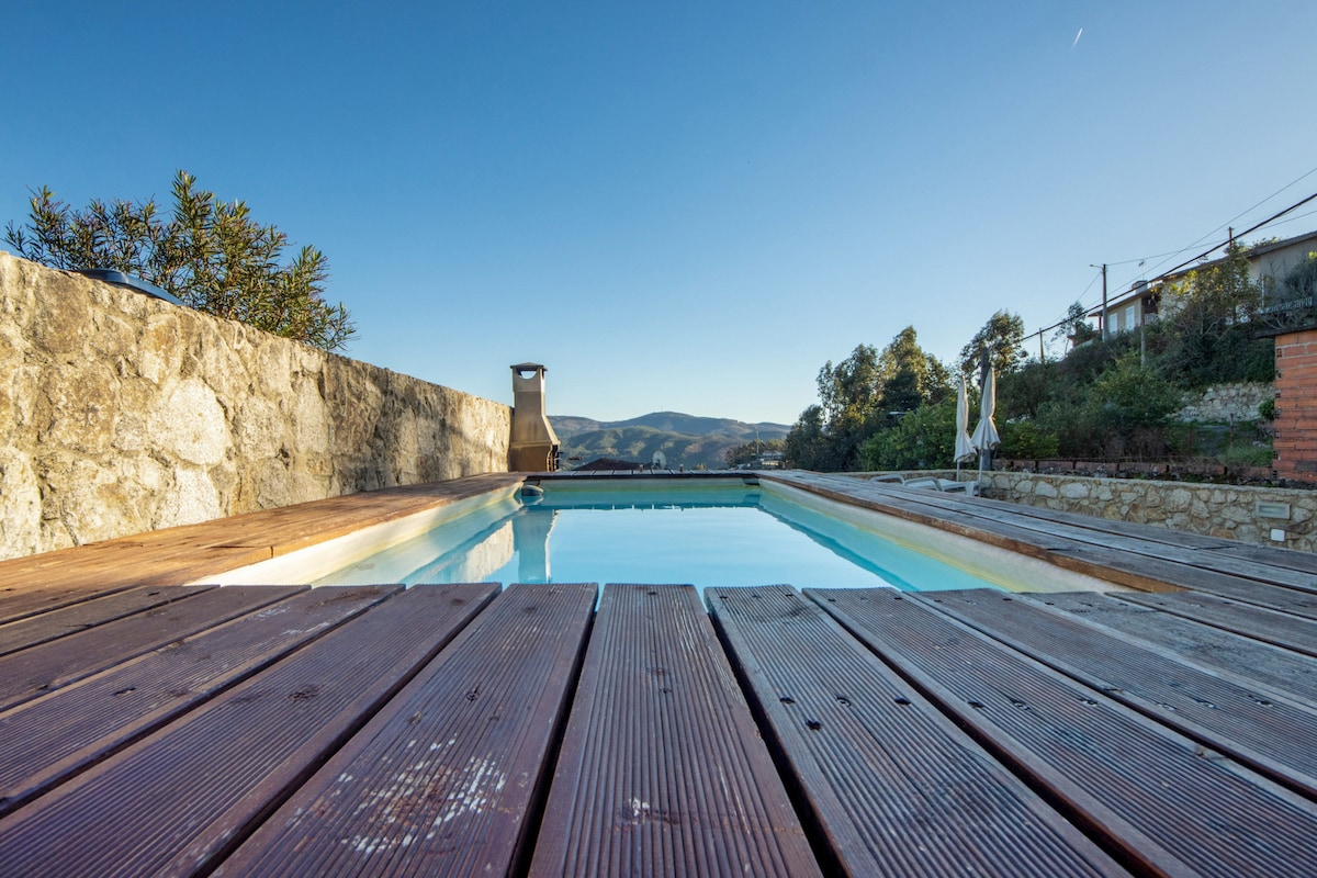 A private outdoor pool, surrounded by wooden decking, is showcased under a clear blue sky. A stone wall provides a natural backdrop, while distant green hills are visible in the background, enhancing the serene outdoor space.
