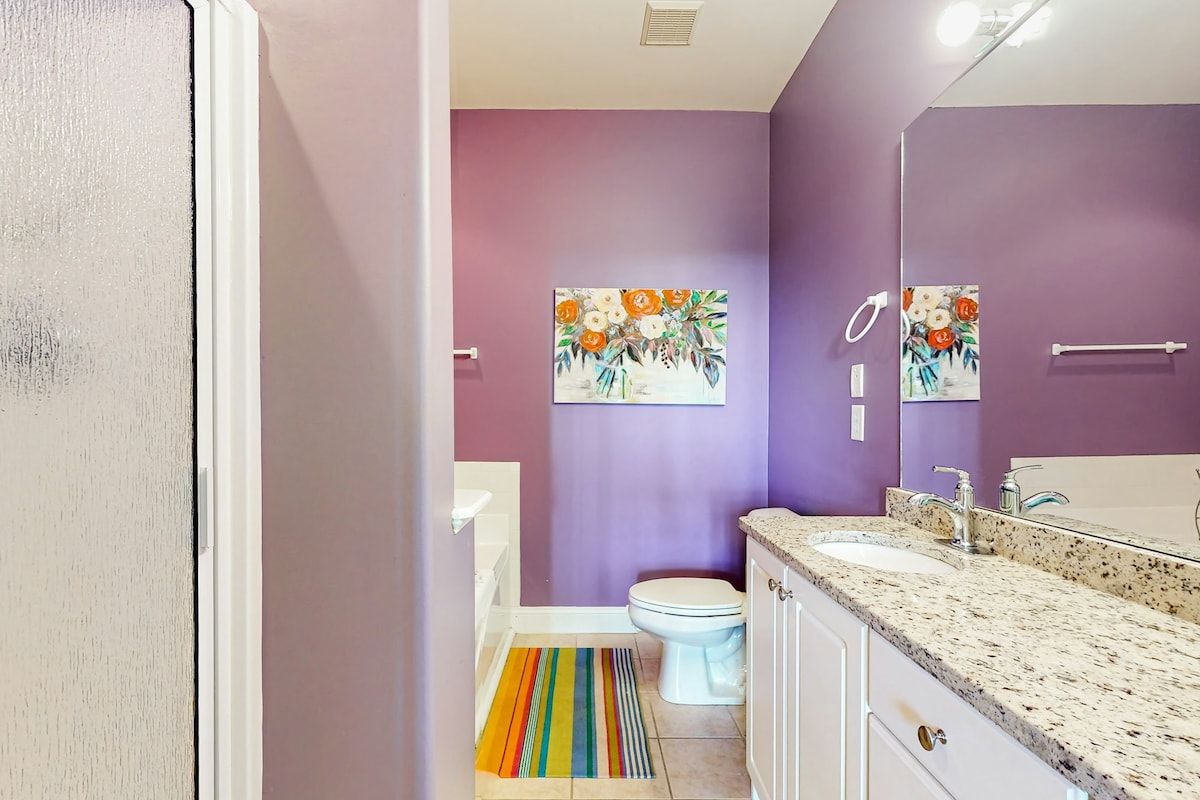 A brightly colored bathroom features a purple accent wall and a large mirror above the vanity. A colorful striped rug is laid across the floor, adding a touch of vibrancy. Modern fixtures and a toilet are positioned against the wall, creating a functional and inviting space.