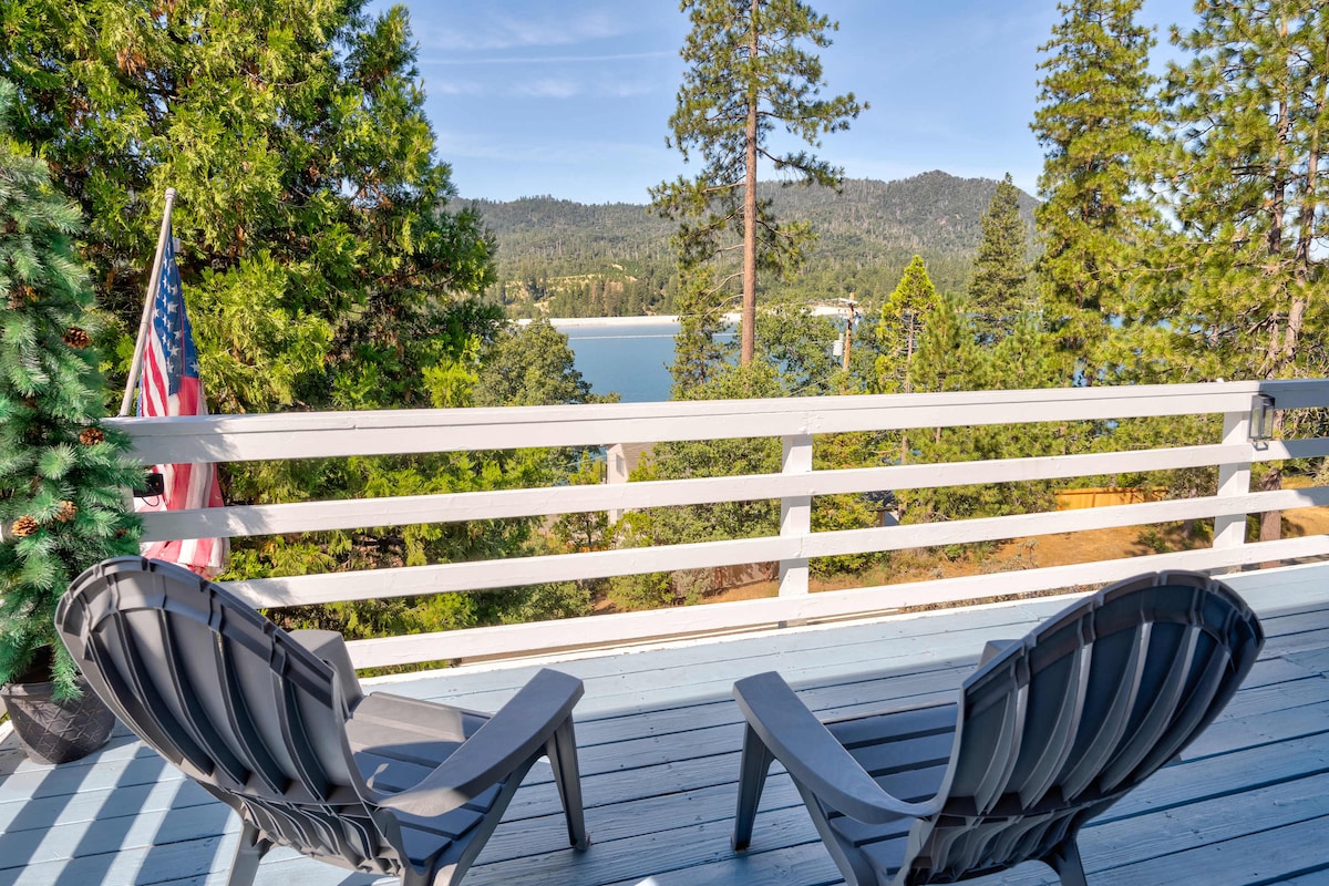 Two outdoor chairs are positioned on a deck overlooking a serene lake surrounded by tall trees and mountain views. A flag is displayed nearby, with clear blue skies enhancing the tranquil atmosphere.