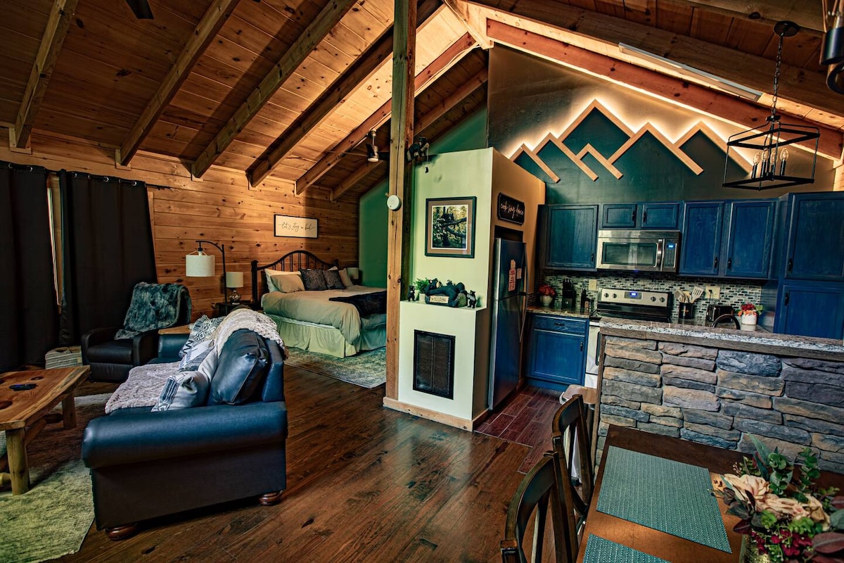 An open living area is characterized by vaulted wooden ceilings and a warm, rustic design. A king bed is positioned near the kitchen, which features blue cabinets and granite countertops. A cozy seating area is complemented by a wooden coffee table, enhancing the cabin's inviting feel.