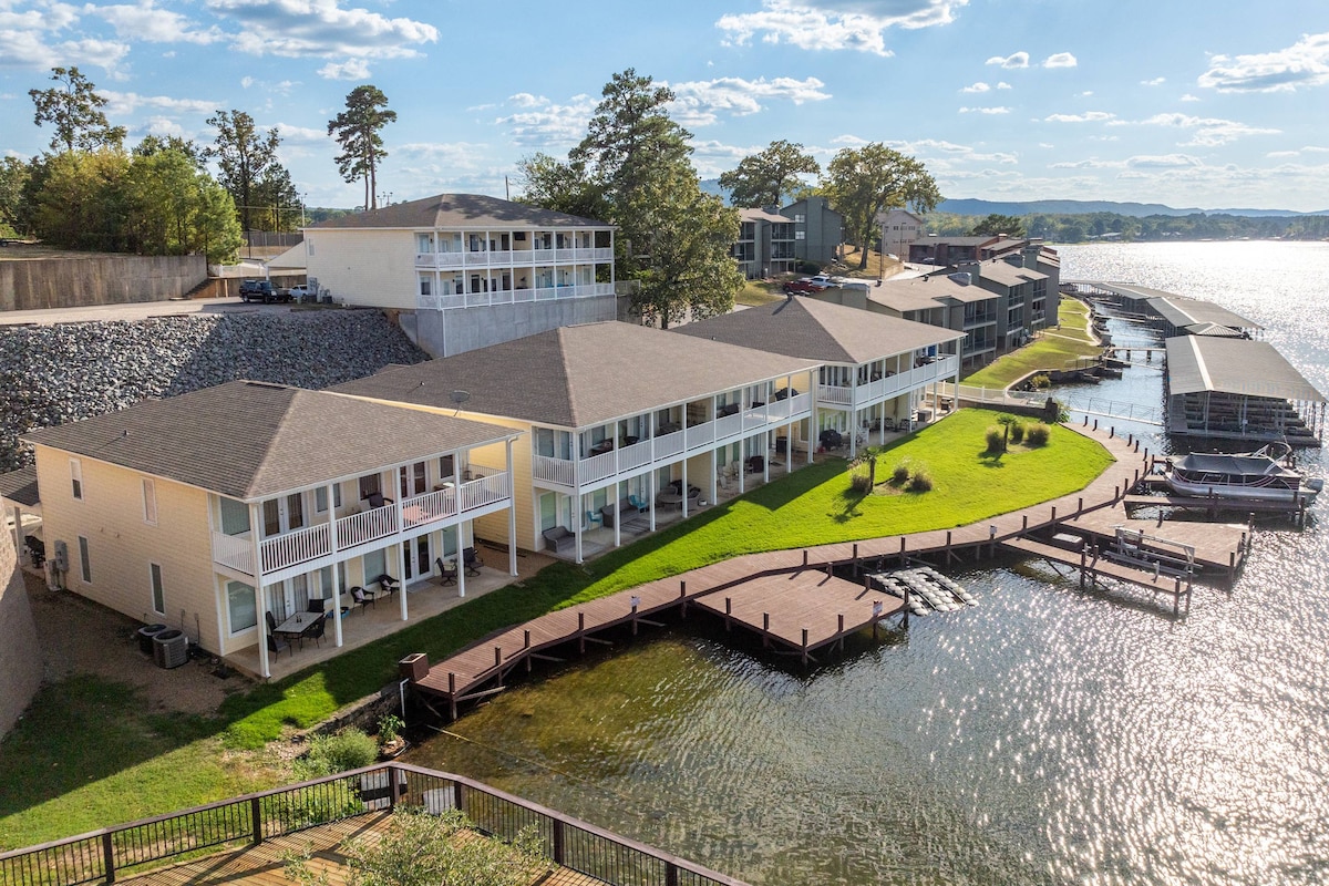 The image shows a waterfront property comprising several connected buildings, surrounded by lush green grass. A wooden dock extends into the water, with boats visible nearby. The scene is brightened by clear blue skies and soft clouds, creating a serene lakeside atmosphere.