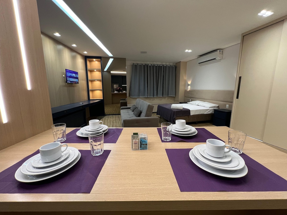 A modern studio layout features a dining area with neatly arranged dishes, cups, and glasses on a light wooden table. A comfortable sleeping area is visible in the background, alongside a cozy lounge space, while the television is mounted on the wall, enhancing the contemporary design.