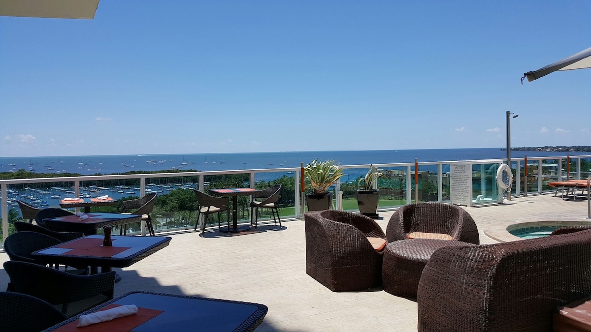 An outdoor terrace features a spacious seating area with wicker furniture, overlooking the water. A panoramic view of the bay is visible in the distance, accompanied by clear blue skies. Tables are arranged for dining, creating an inviting atmosphere for relaxation.