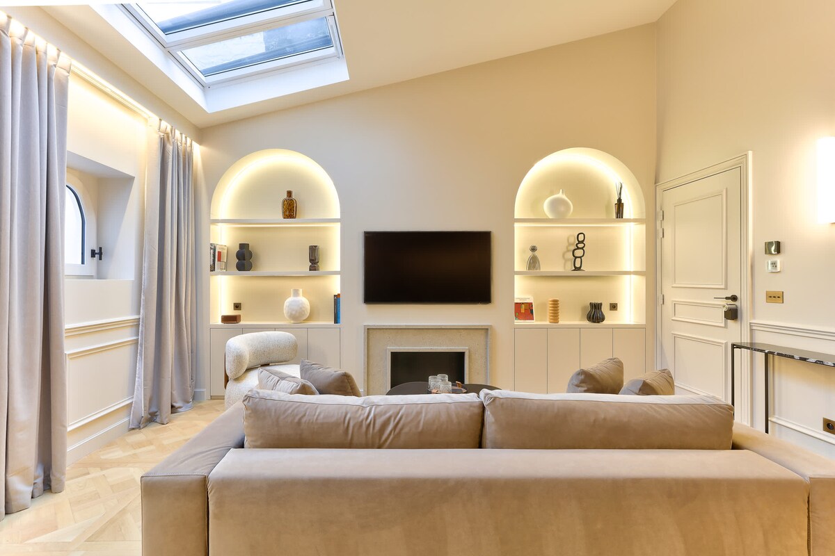 The living room features a light color palette with soft beige furnishings and a modern sofa. A mounted TV is centered above a sleek stone fireplace. Built-in shelves display decorative items, with recessed lighting providing a warm glow, while large windows and a skylight allow natural light to fill the space.
