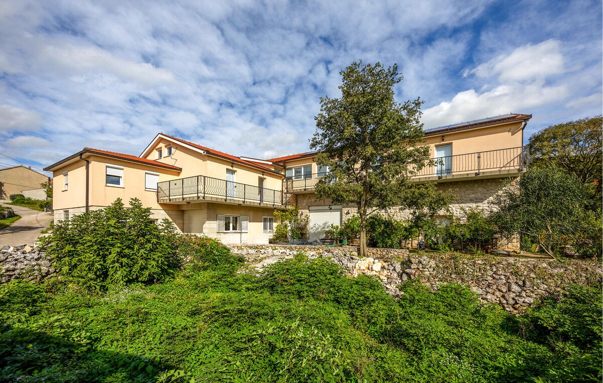 The modern building is set against a backdrop of green foliage and stone walls. A balcony runs along the upper level, offering views of the surrounding area. The structure features large windows that allow natural light to illuminate the interior spaces.