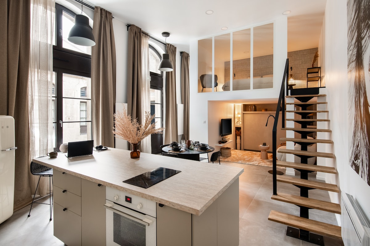 An open-plan living area showcases a modern kitchen island with a stovetop and seating. Large windows allow natural light to fill the space, complemented by elegant curtains. A staircase leads to a mezzanine level, while a cozy living area with soft furnishings provides comfort and style.