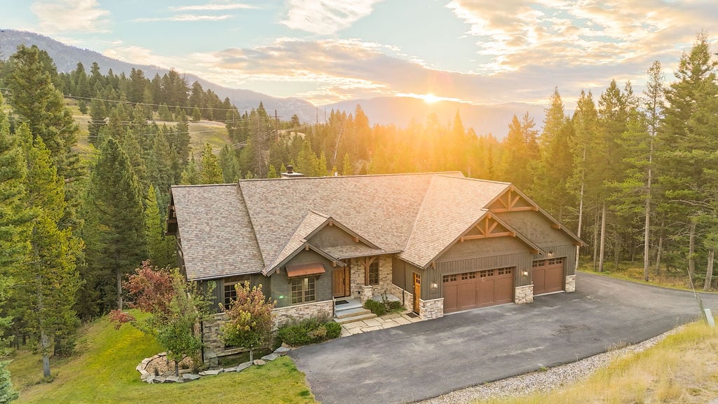 The exterior of a spacious luxury home is showcased, featuring a combination of wood and stone materials. A driveway leads to a three-car garage. Sunlight casts a warm glow against the backdrop of trees and mountainous terrain, promoting a serene atmosphere.