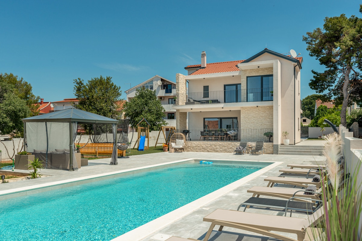 A modern villa showcases a spacious exterior with a private heated pool surrounded by lounge chairs. A covered seating area is positioned nearby, along with a children's playground visible in the serene courtyard setting. The structure features an appealing multi-level design with balconies.