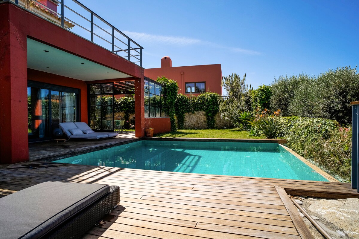A spacious outdoor area features a private swimming pool surrounded by wooden decking. Comfortable loungers are positioned nearby. The villa's large glass doors reflect ample sunlight and offer views of the lush garden and surrounding greenery, creating a serene atmosphere.