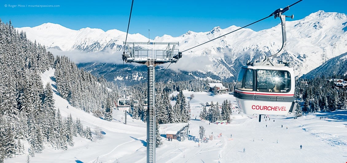 A gondola labeled 'Courchevel' is suspended above a snowy landscape, with pristine white slopes and evergreen trees surrounding it. In the background, majestic mountains are partially obscured by clouds, creating a serene winter scene.