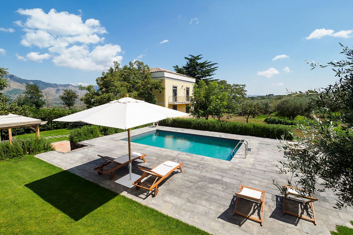 An outdoor area features a private swimming pool surrounded by a spacious stone patio. Sun loungers are positioned under an umbrella, providing shade. Lush greenery and trees extend into the distance, offering a serene view of the surrounding landscape and nearby hills.