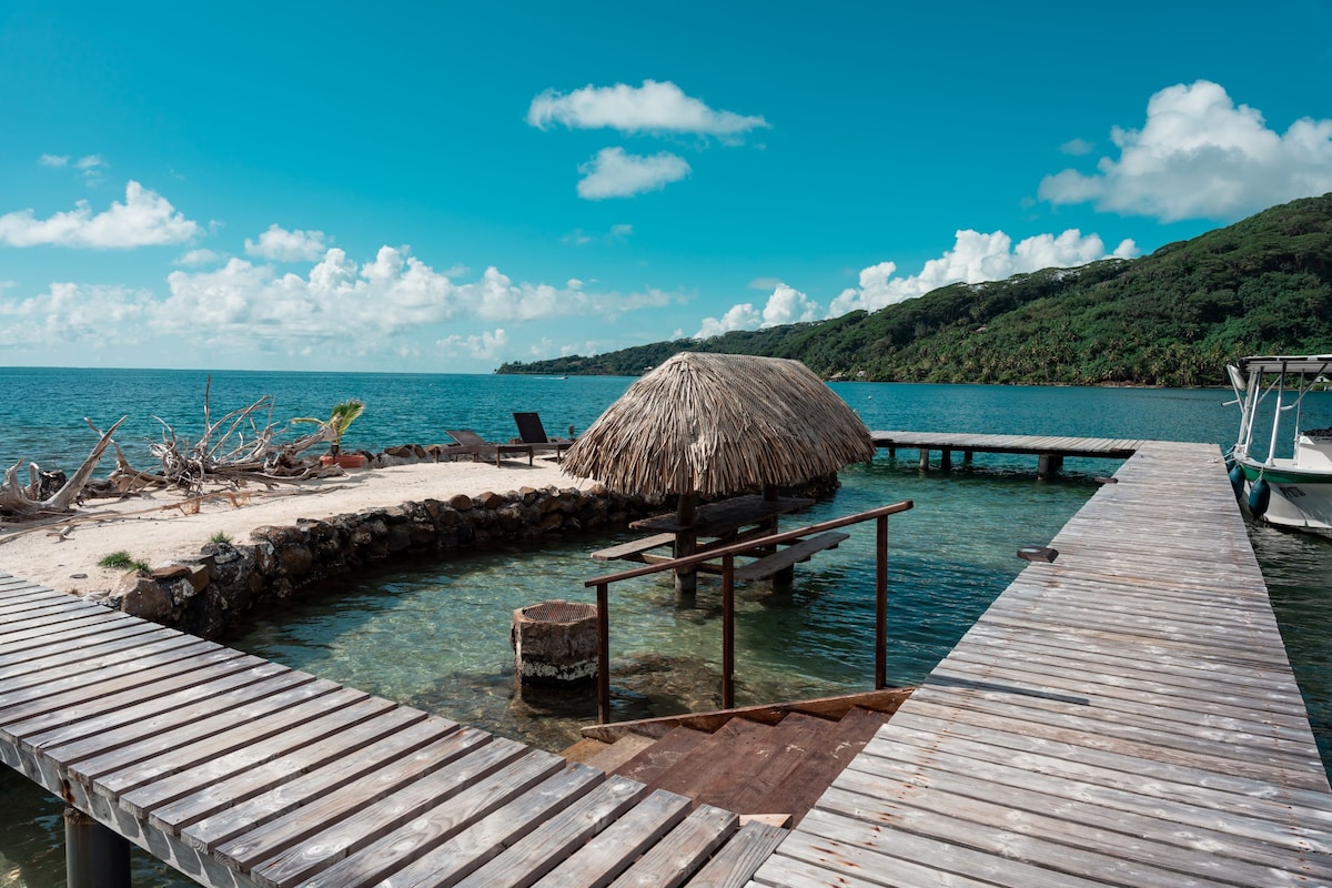 A private pontoon extends over clear turquoise waters, featuring a thatched-roof hut and wooden decking. Sun loungers are positioned for relaxation, with the backdrop of a lush green hillside and scattered clouds in the bright sky.