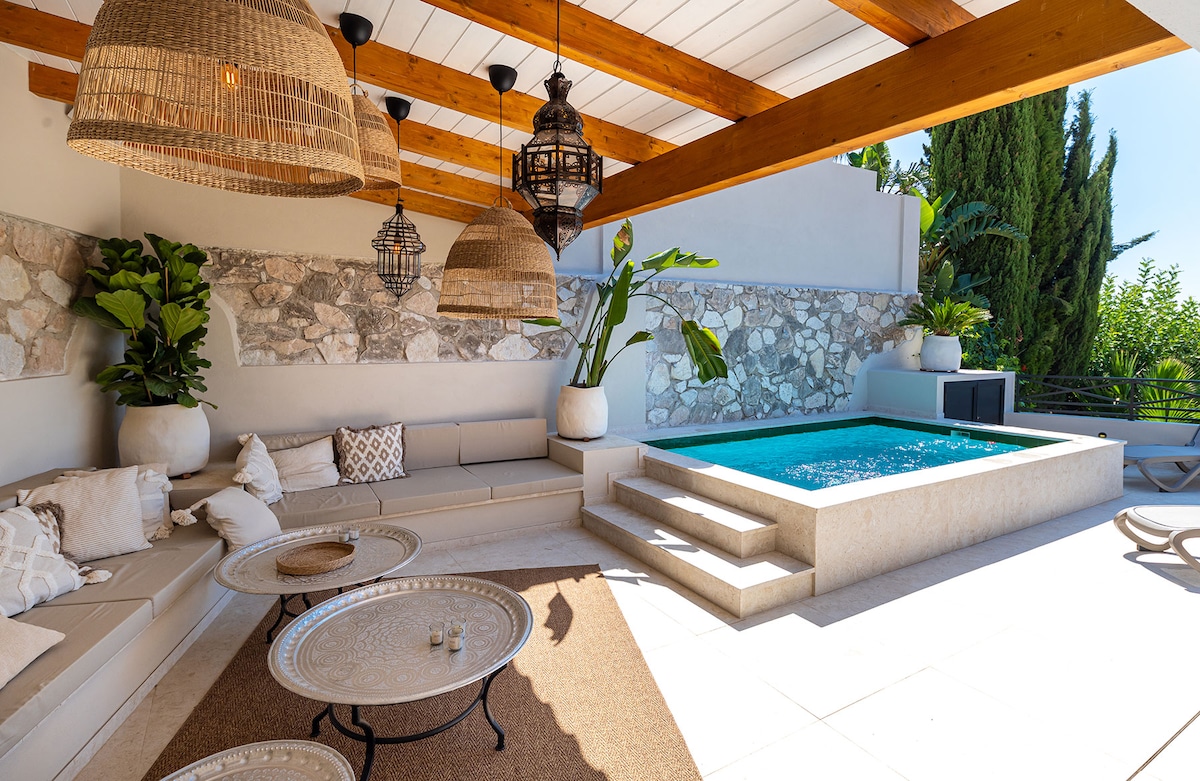 An outdoor seating area features a comfortable, U-shaped sectional sofa adorned with neutral cushions. A small, circular table is placed in front of the sofa, complementing the space. Overhead, woven pendant lights hang from a wooden beam, while a private dipping pool is situated nearby, surrounded by lush greenery.