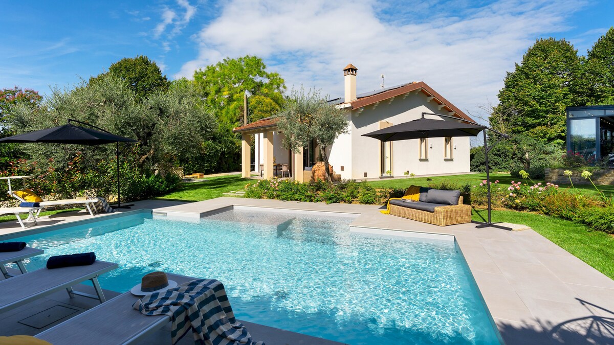 The swimming pool is surrounded by a spacious deck featuring sunbeds and adjustable umbrellas. The villa is visible in the background, set against a lush garden with greenery and colorful plants, creating a relaxing outdoor environment.