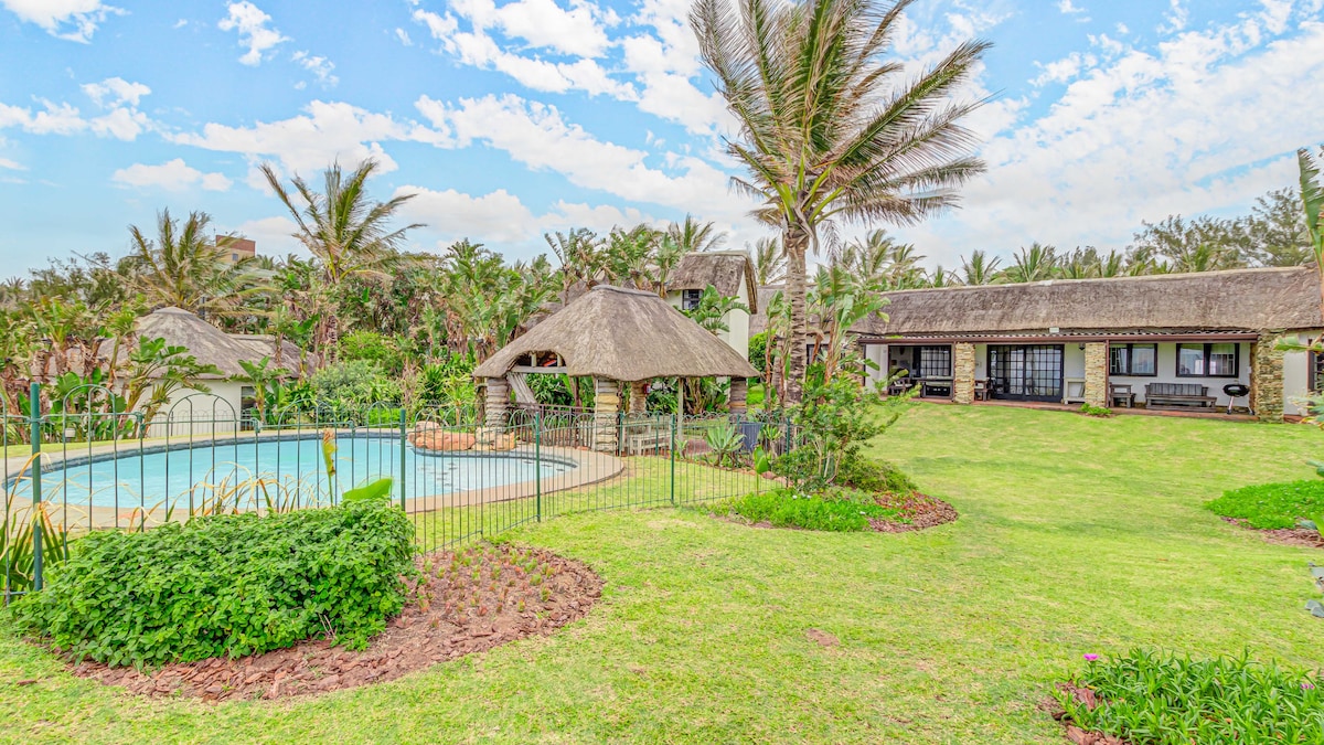 The outdoor area features a refreshing swimming pool surrounded by lush greenery and palm trees. A traditional thatched-roof gazebo is visible nearby, providing a shaded seating area. The expansive lawn offers ample space for relaxation and outdoor activities, enhancing the tranquil ambiance of the property.