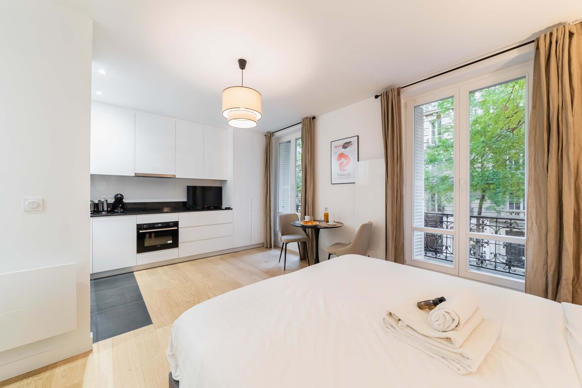 A well-lit studio features a comfortable bed in the foreground. A small dining area is seen with two chairs and a table. The kitchen is modern and equipped, with dark countertops and white cabinetry. Large windows offer views of greenery outside.