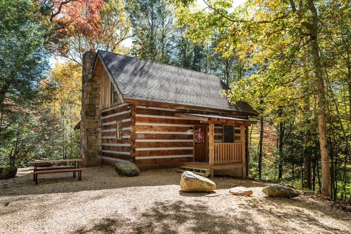 A charming log cabin is set amidst a vibrant forest, with colorful foliage surrounding the structure. The gravel pathway leads to the entrance, featuring a welcoming porch and a natural stone facade. Large boulders are placed decoratively in the yard, enhancing the rustic appeal.