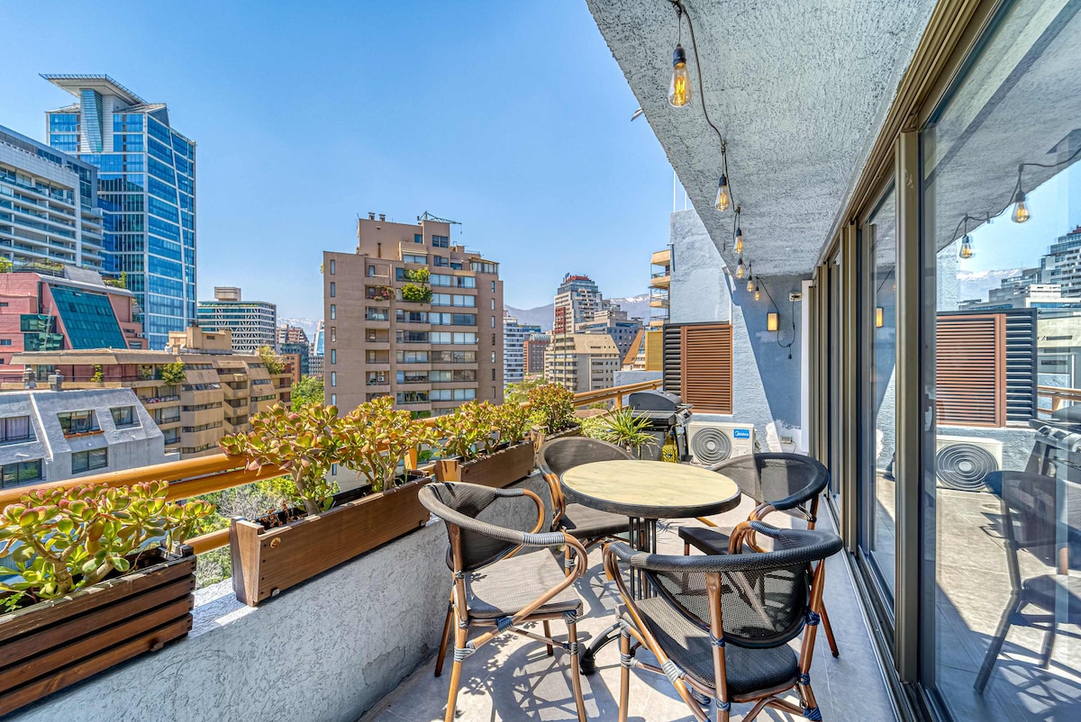 A spacious terrace offers city views, framed by potted plants. Outdoor seating is provided with a round table and four chairs, creating an inviting area for relaxation or dining. Clear skies and nearby buildings are visible in the background.