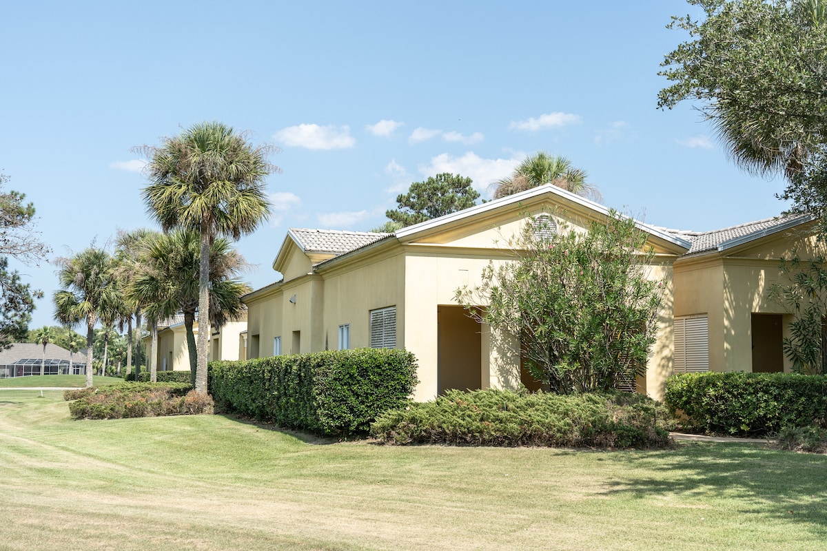 A row of villas is showcased against a clear blue sky, with palm trees and manicured green lawns framing the scene. Each villa features a covered entrance, and shrubs provide a natural boundary, contributing to a serene, inviting environment.